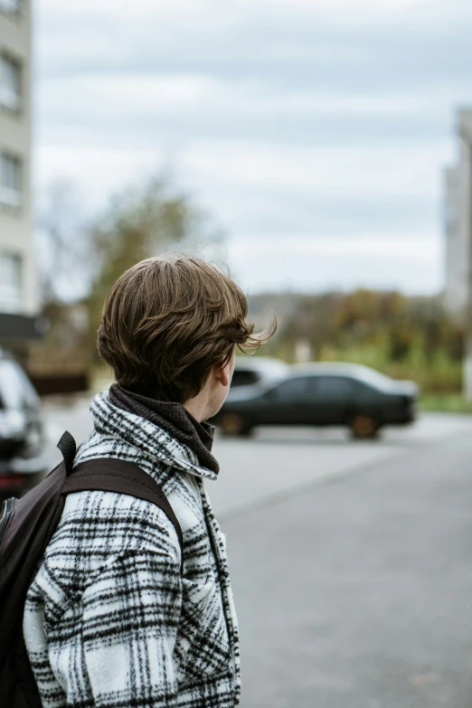 an older woman with short hair and a jacket on stands by a street and stares out to the distance