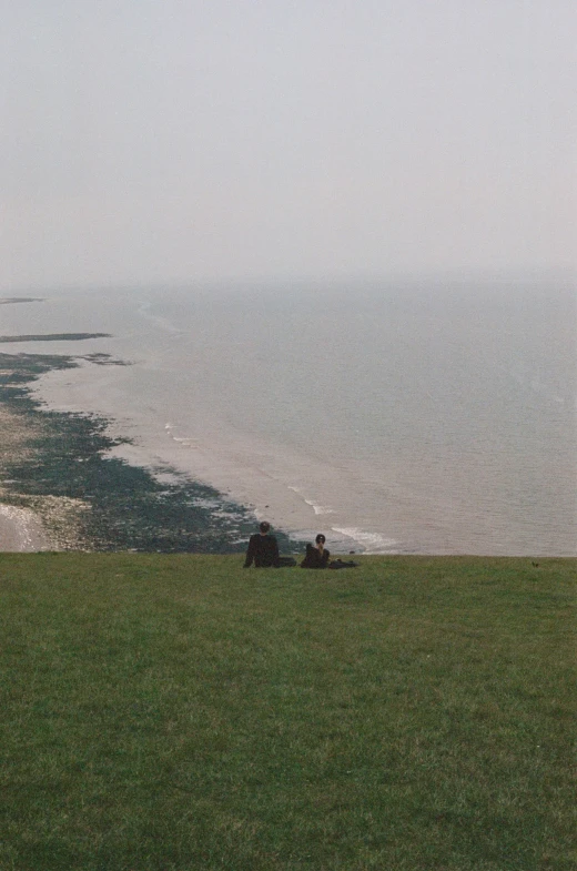 two people sitting on a grassy hill near a body of water