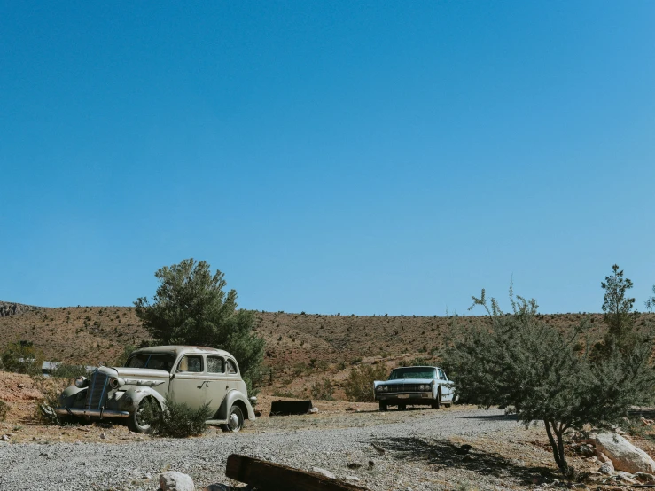 two old cars parked in the desert, next to trees and a bench