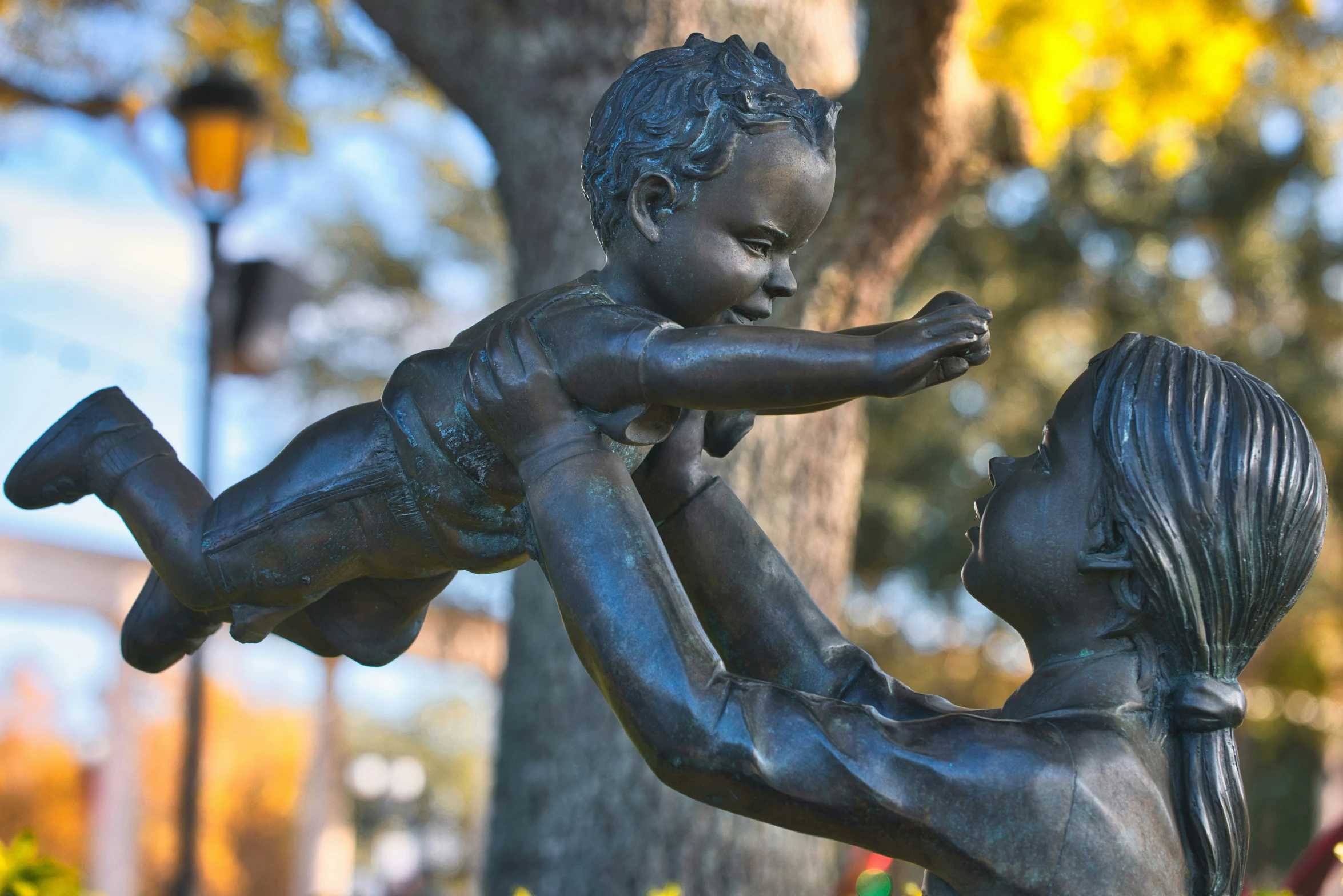 a bronze statue of a child holding an infant standing next to a tree