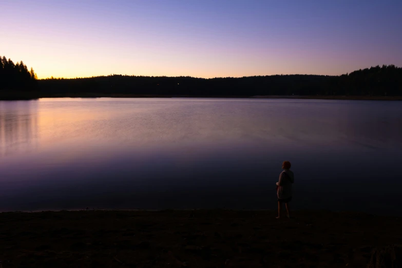 a man standing on a shore line by a lake at sunset