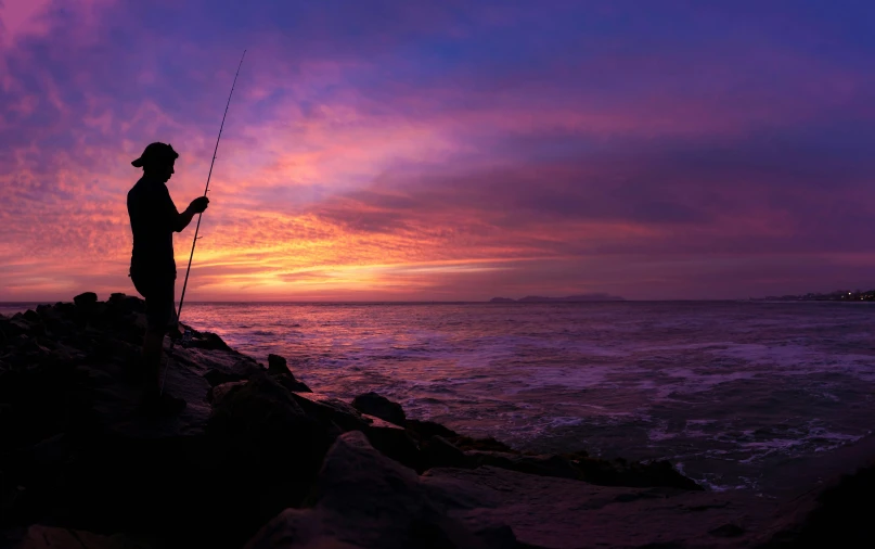 a man standing on top of a rock next to the ocean while holding a fishing pole