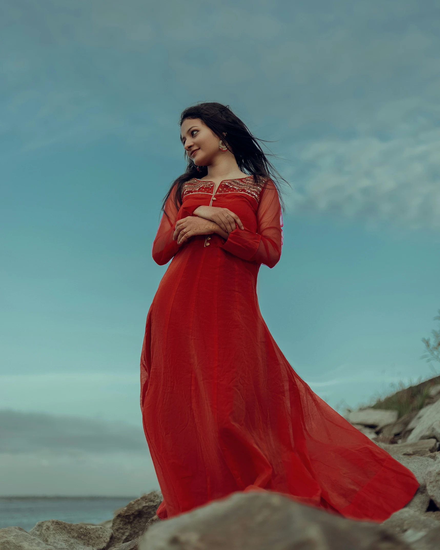 a woman wearing a red dress stands on a rocky shoreline