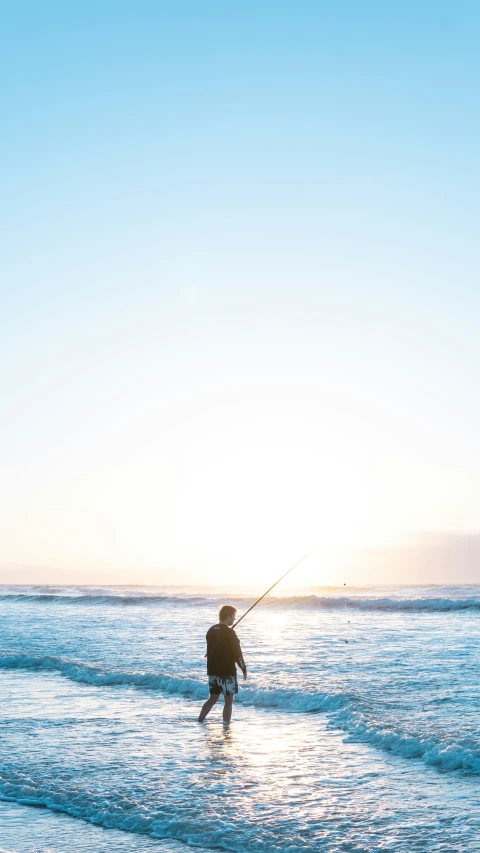 a man is wading in the shallows of the ocean, pulling his fishing rod up