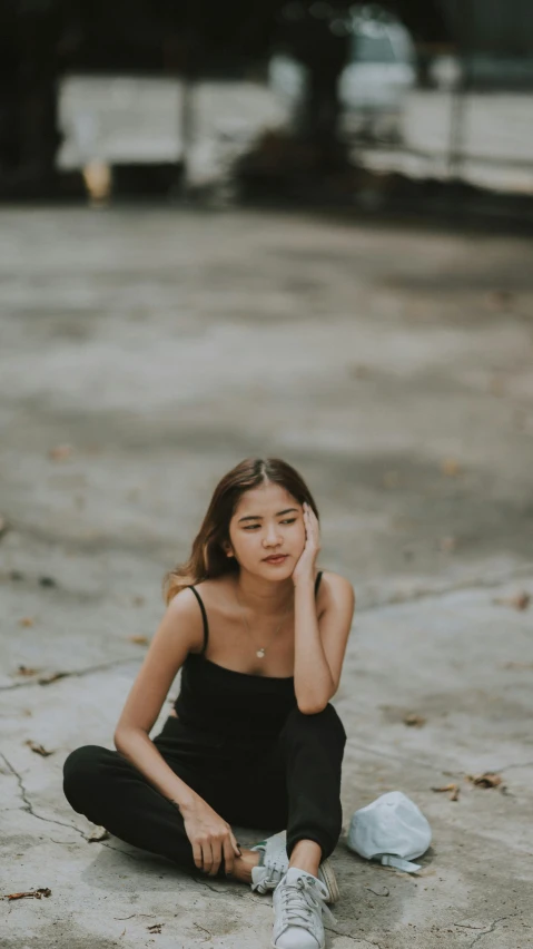 a young lady with dark hair sits on the concrete and holds her head to her side