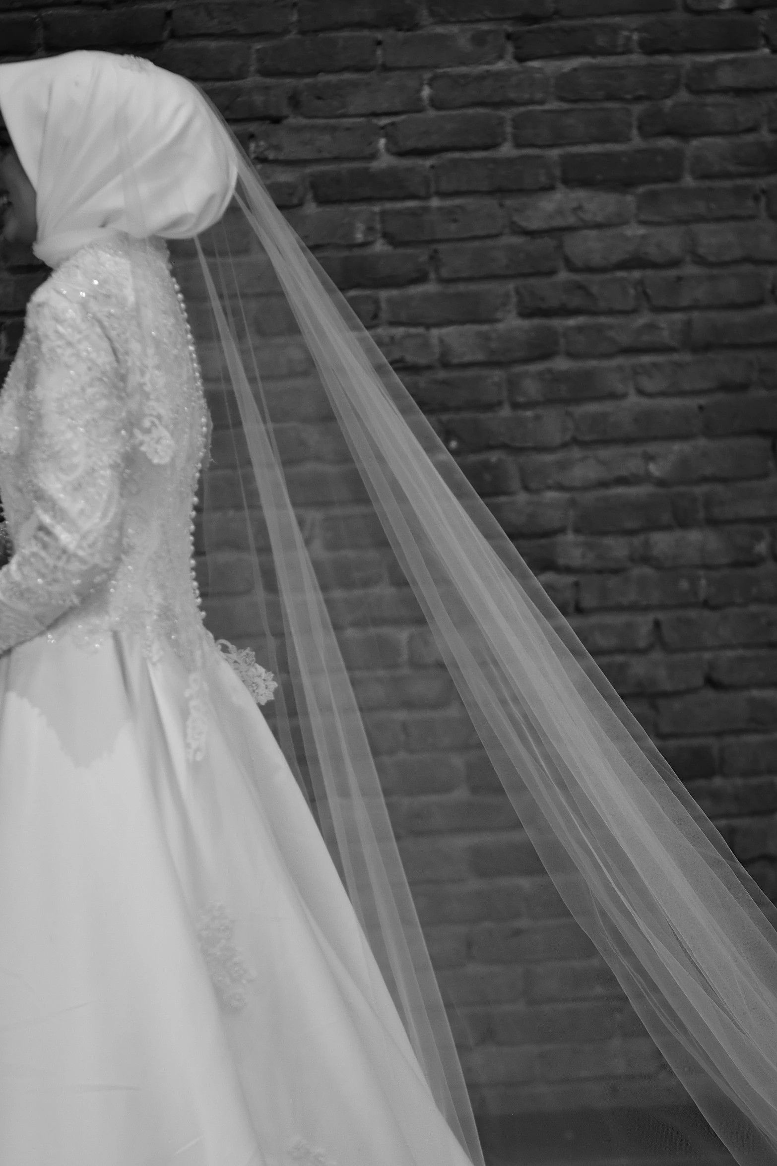 bride and groom standing by brick wall with veil blowing in wind