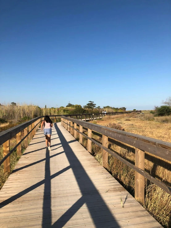 a woman walking down a long, wooden bridge across a marsh