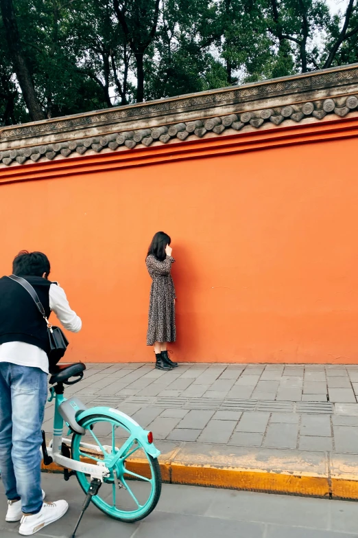 two people on street corner near a bicycle leaning against orange wall