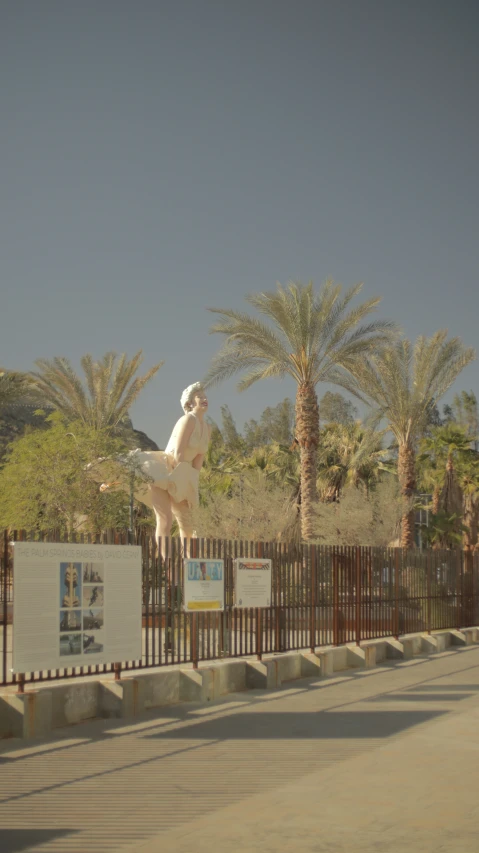 an art exhibit of a dog standing on its hind legs in the foreground with palm trees behind it and a brown fence with signs below it