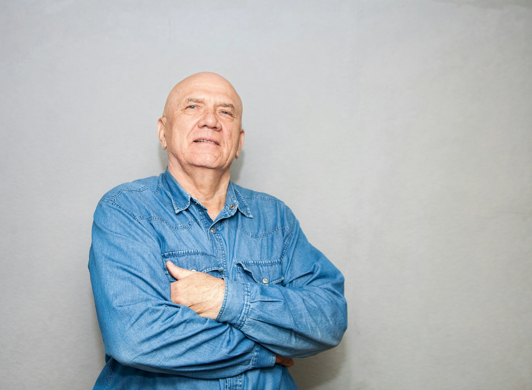 an elderly man with his arms crossed standing against a grey wall