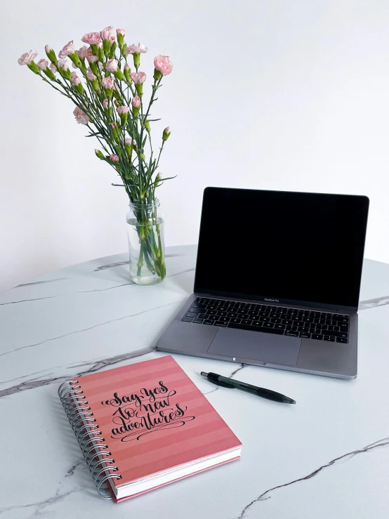 an open notebook sits next to a vase of flowers on a marble table