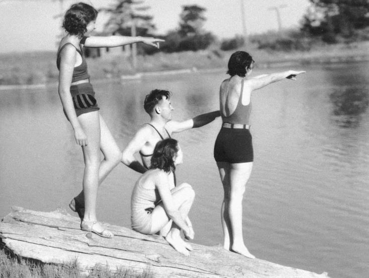 three young women standing and sitting on a jetty with one pointing at soing