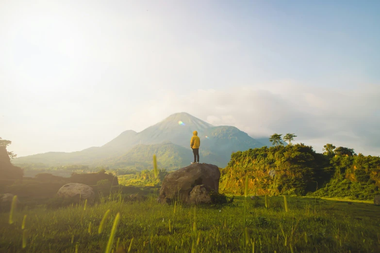 man stands on large rock in grassy field with rocky terrain and mountain in background