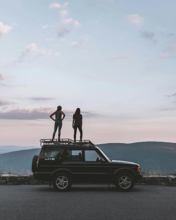 two people stand on top of their suv in the mountains