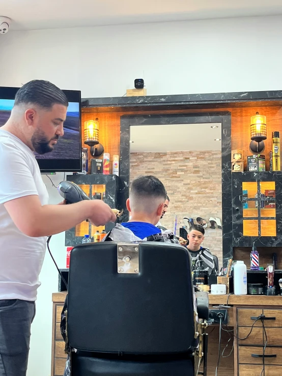 a man sitting in a barber chair and another barber  his hair