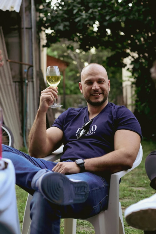 a man in a blue shirt sitting on a white chair holding up a wine glass