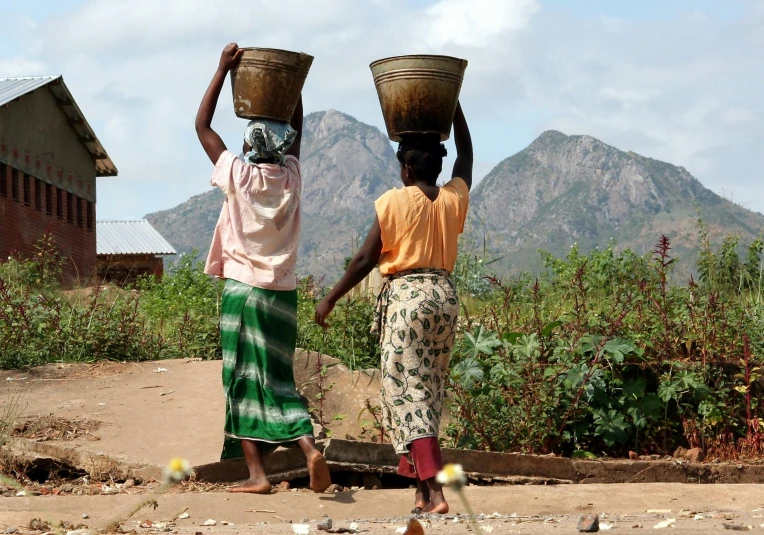 two women carrying water on their head walking down a dirt road