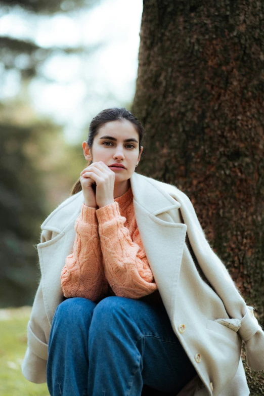 a woman wearing a cream coat sitting under a tree