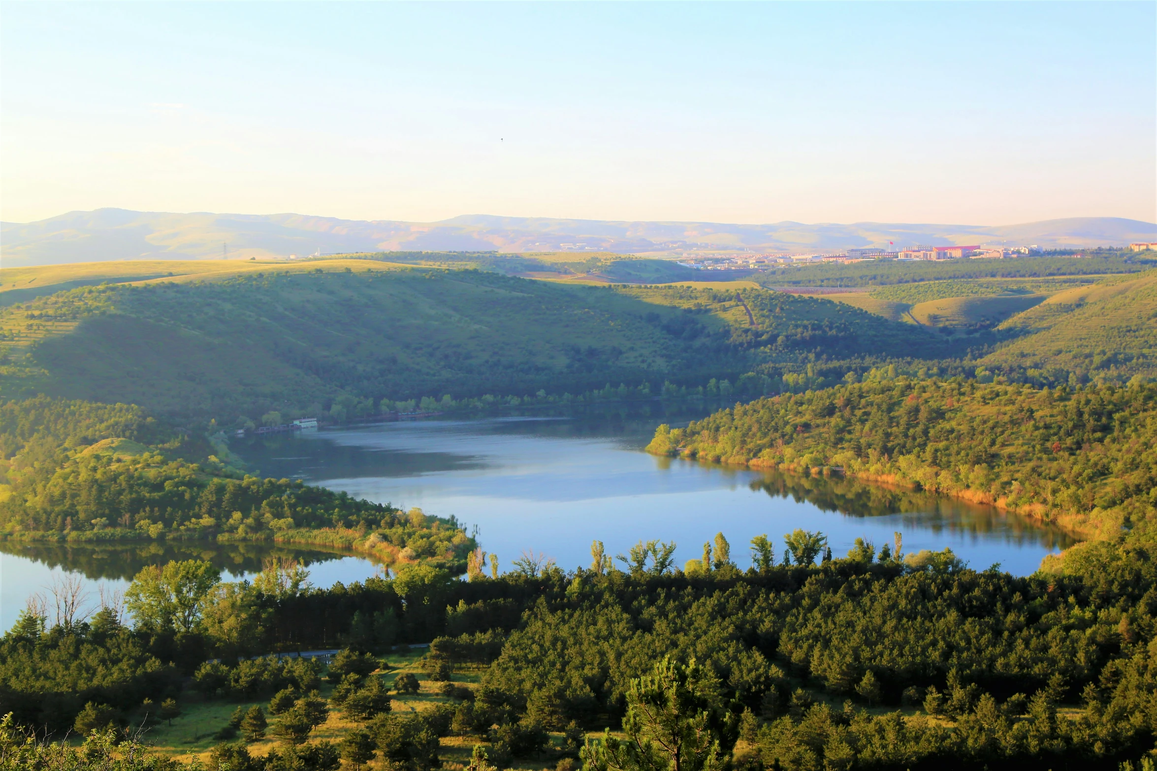 a lake in the middle of green hills with trees and some buildings on the other side