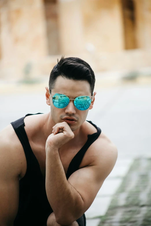 a young man is leaning against the side of a brick wall wearing a tank top and blue mirrored sunglasses