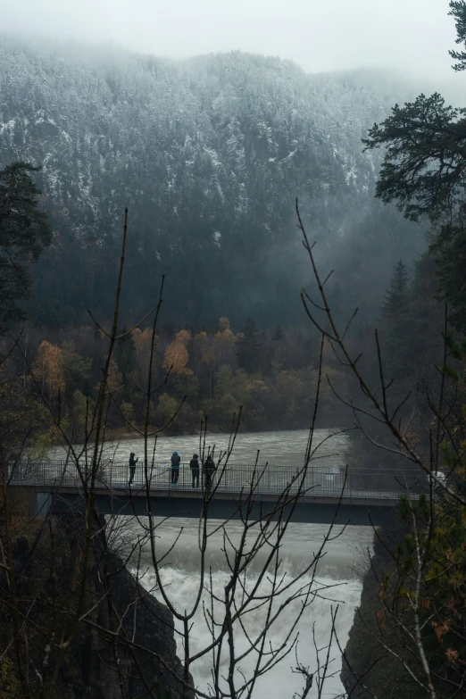 people stand on a bridge over a stream, near a mountain