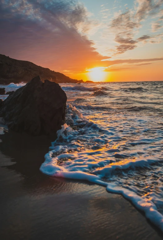 a sunset with a wave coming in towards the shore and the rocks are just starting to fade