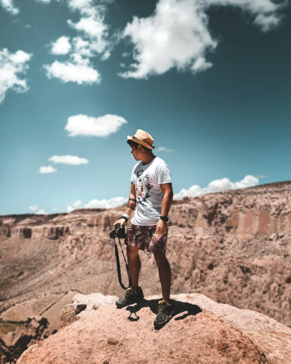 a man with a hat and glasses stands on top of a rock, while holding a camera