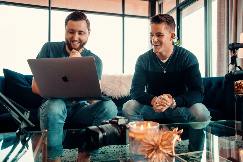 two men sitting on a couch in front of a laptop