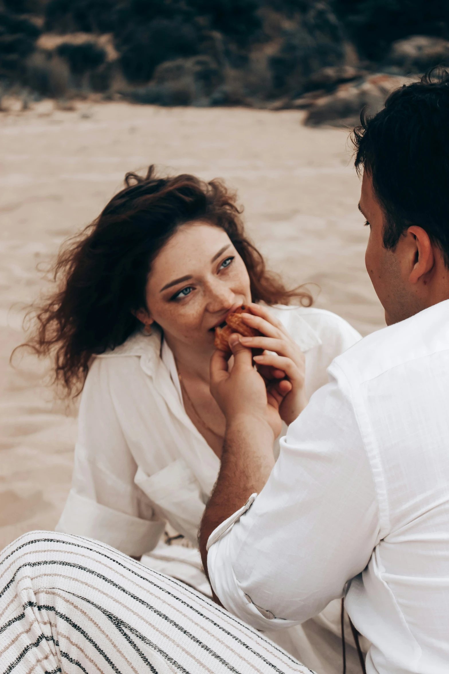 two people sitting on the sand, one of them eating soing
