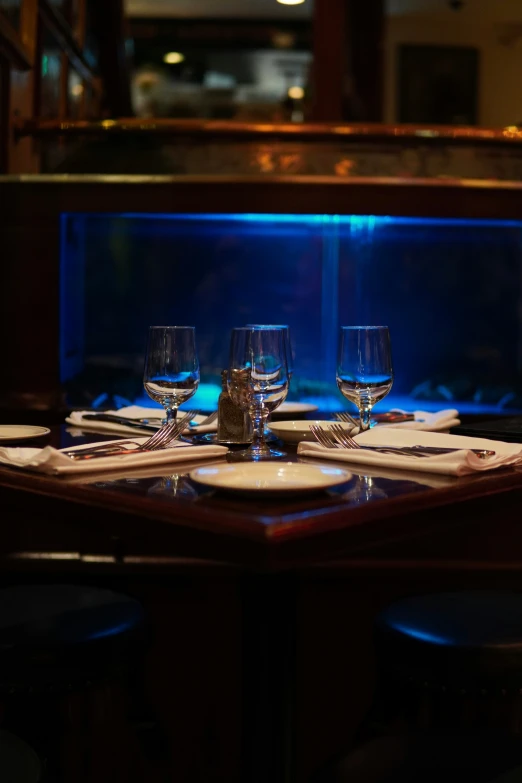 a dining room table with three glasses with lit blue fish tanks in the background