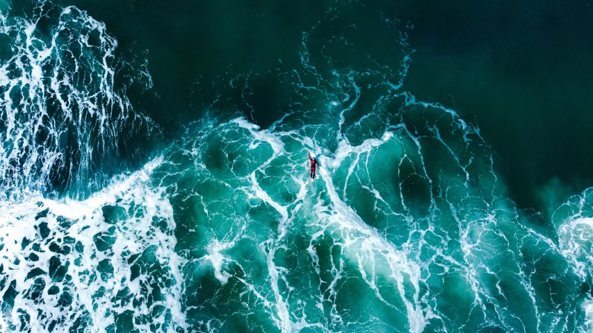 the aerial view of a man in a red jacket standing in water on a surfboard