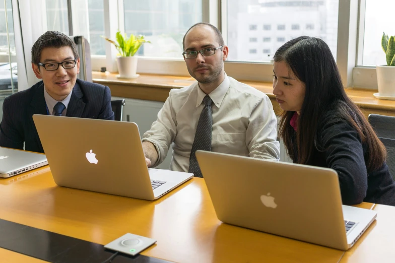 three people in business suits sitting at a desk looking at laptops