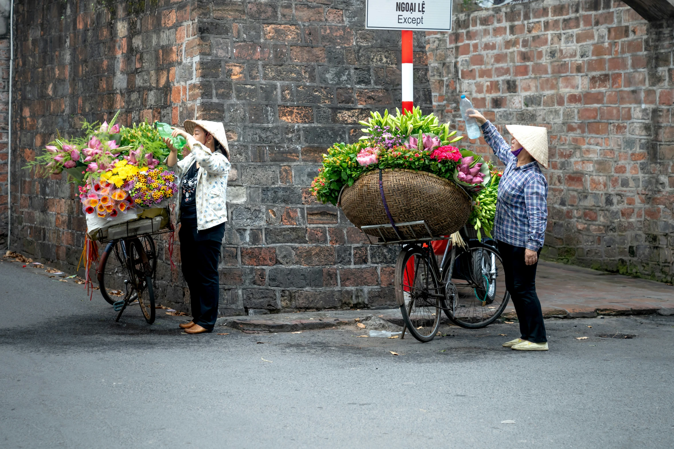 two women standing next to a brick wall with flowers