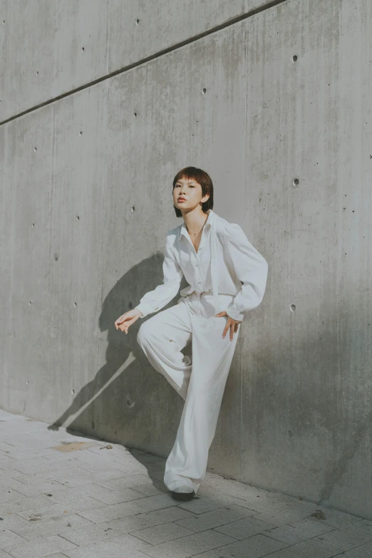 a woman wearing white pants and shirt leaning against a concrete wall