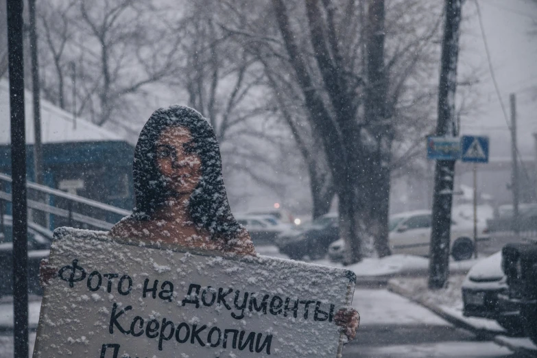 snow falls on a woman holding a sign in the street