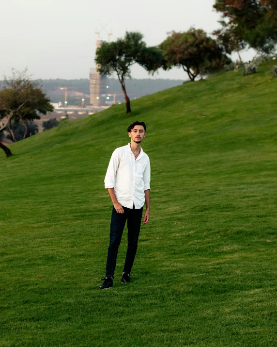 young man standing on grassy hill near small lake and buildings