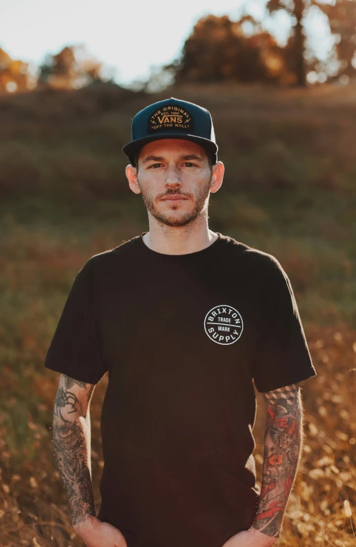 a man with tattoos wearing a black shirt and cap standing in a field