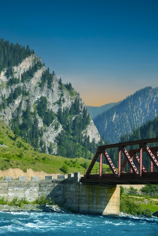 a bridge near a river has a red metal framework and mountain behind it