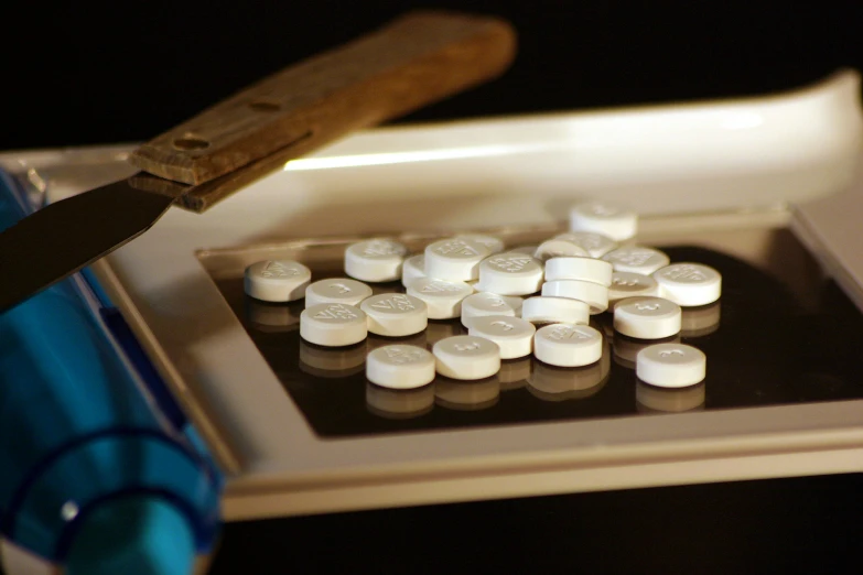 some pills are in a square container while a pair of tongs lays next to them