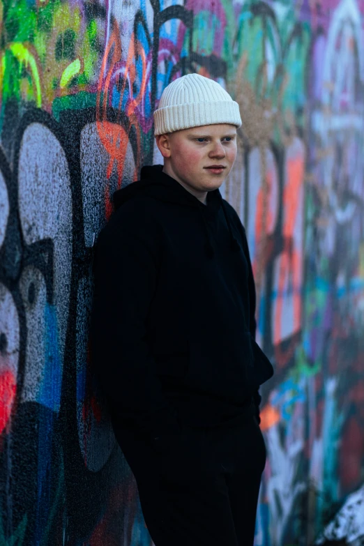 a young man with a hat poses against a wall with graffiti