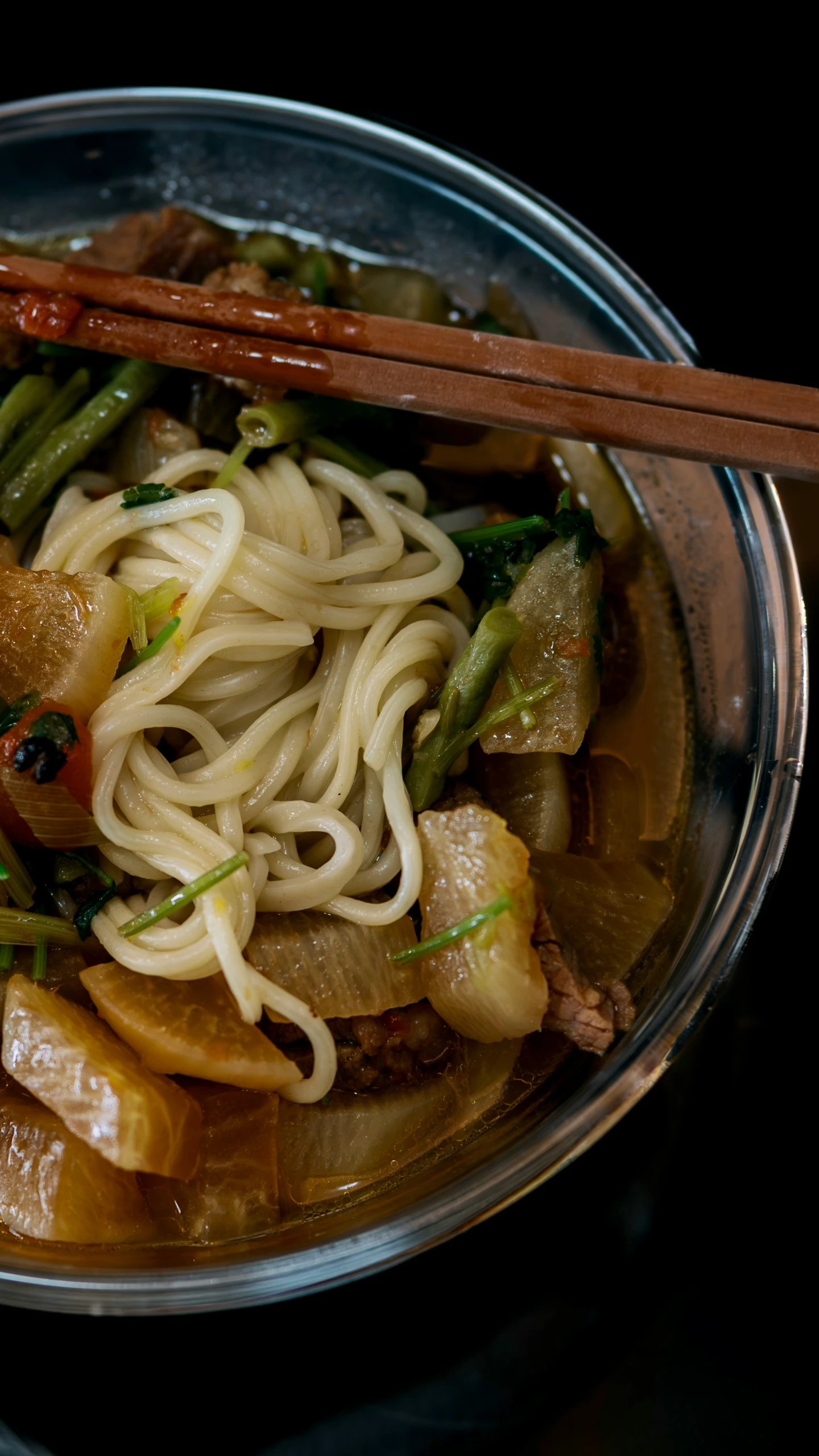 a clear bowl of some noodles and vegetables with chopsticks