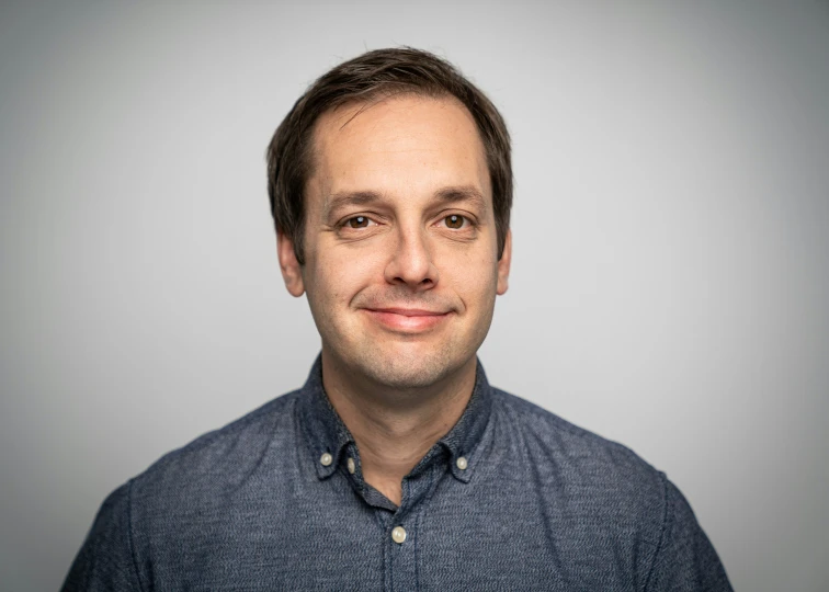 man in blue shirt smiles with gray wall behind him