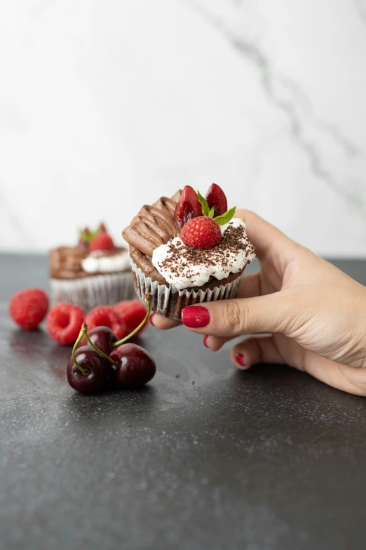 a person holding out a chocolate cupcake with whipped cream, cherries and strawberries