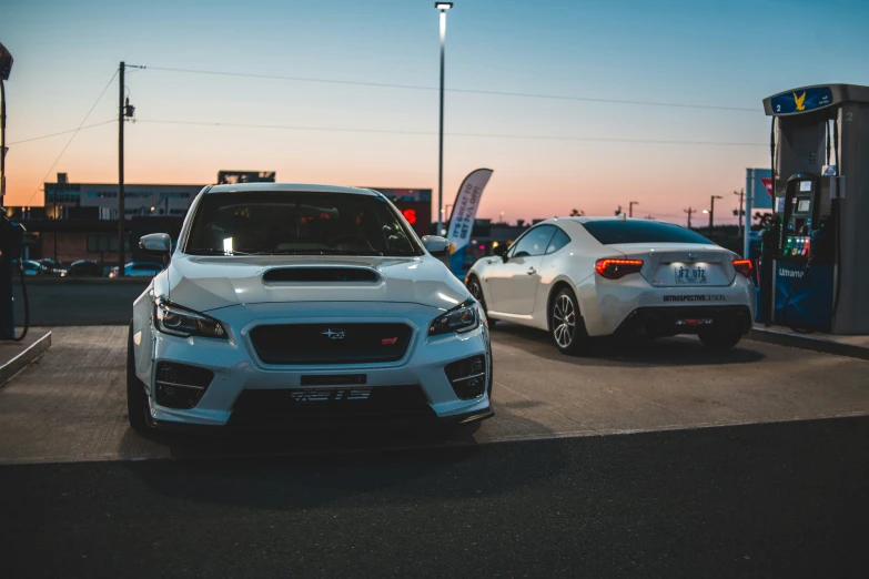 two subaruents, a white sedan and a grey one at a gas station