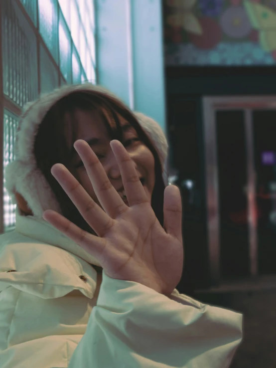 a woman in a white winter jacket has her hand up while covering her face with both hands