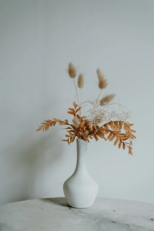 a white vase with dry plants in it on a table